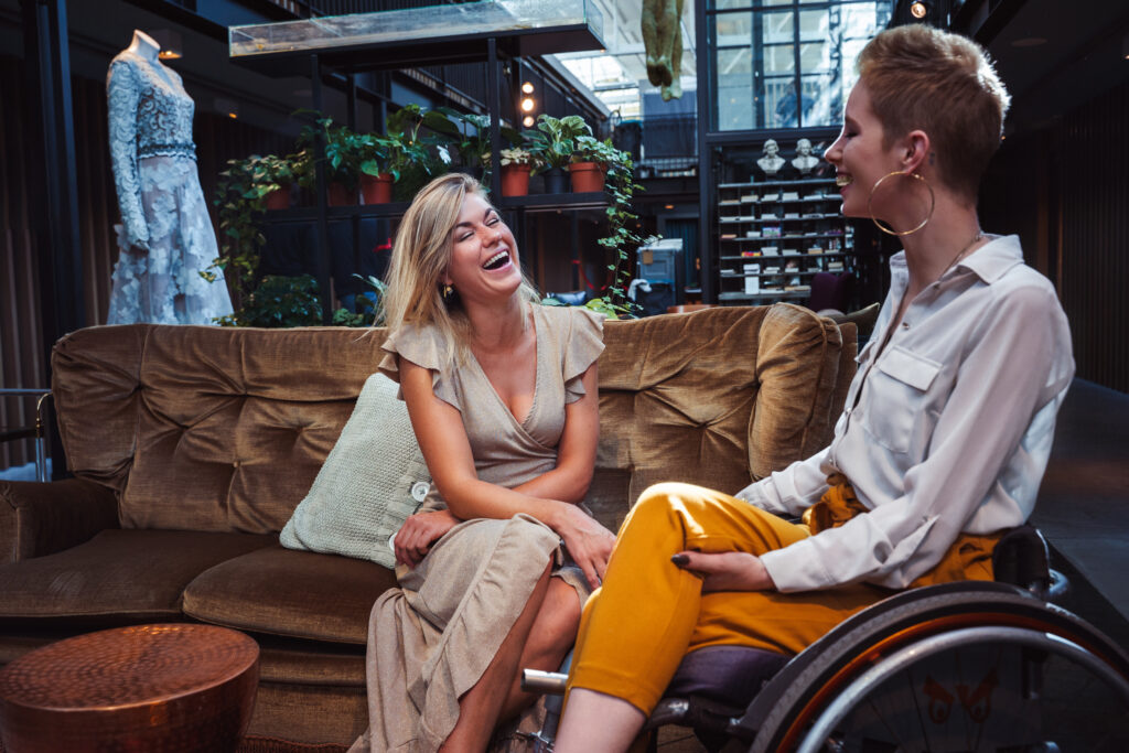 Foto van twee lachende jonge vrouwen, 1 op een bank, 1 in een rolstoel