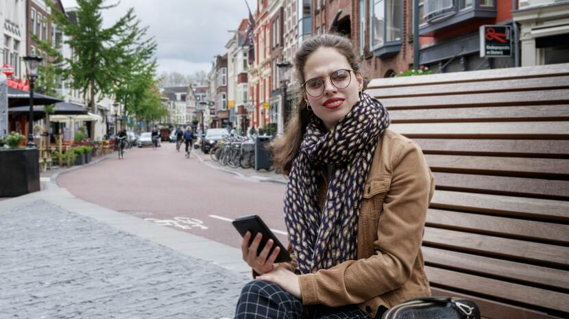 Foto van een jonge vrouw, zittend op een bankje, ze draagt een bruin leren jack en een grote sjaal.