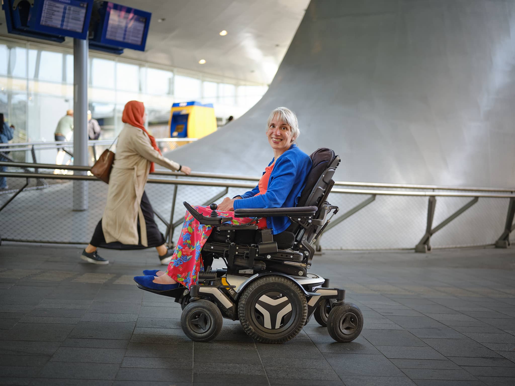 Een vrouw met een blauwe trui en een rode broek zittend in een elektrische rolstoel.