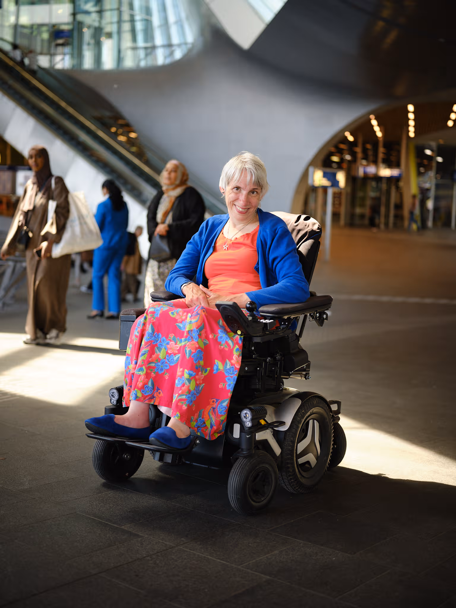 Een vrouw met grijs haar en een lach op haar gezicht zitten in een elektrische rolstoel. Ze kijkt in de camera en heeft een blauw vest en een rood gebloemde broek aan. 