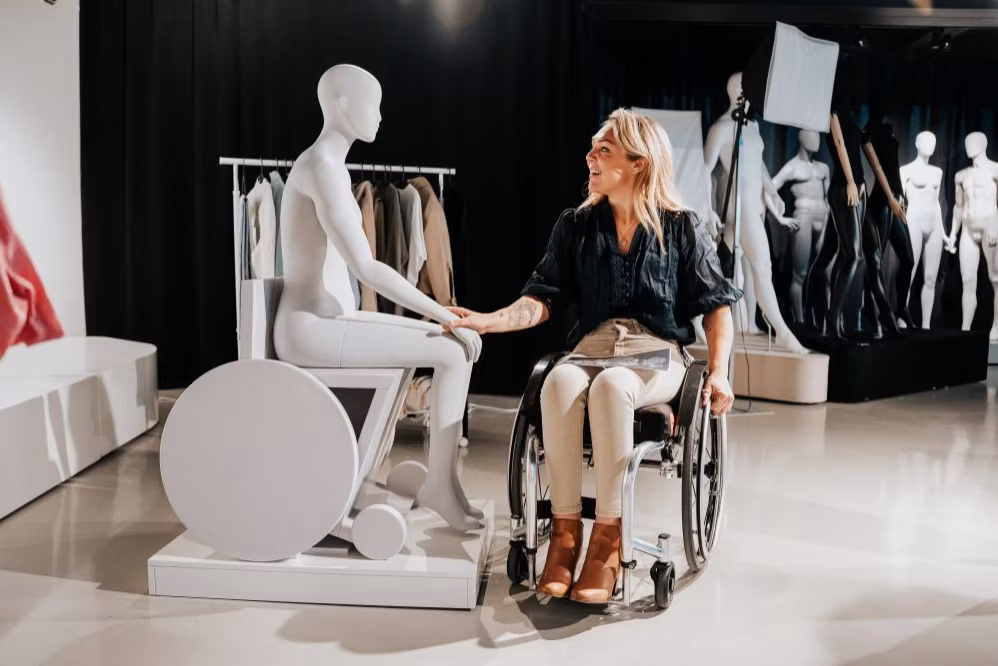 Rechts een blonde dame in een rolstoel, zij geeft een hand aan een geheel witte etalagepop die in een rolstoel zit.