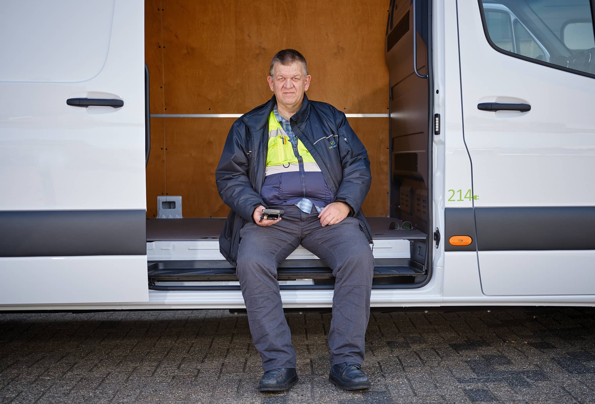 Foto van een man in een blauw pak zittend in de opening van een busje. Hij kijkt recht de camera in. 