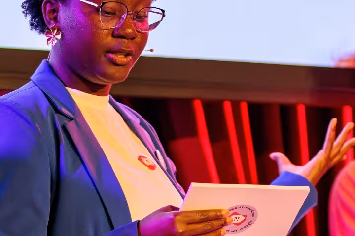 Foto van een jonge dame, ze draagt een bril, een donker colbert en een wit T-shirt. Ze heeft een wit papiertje in haar hand,