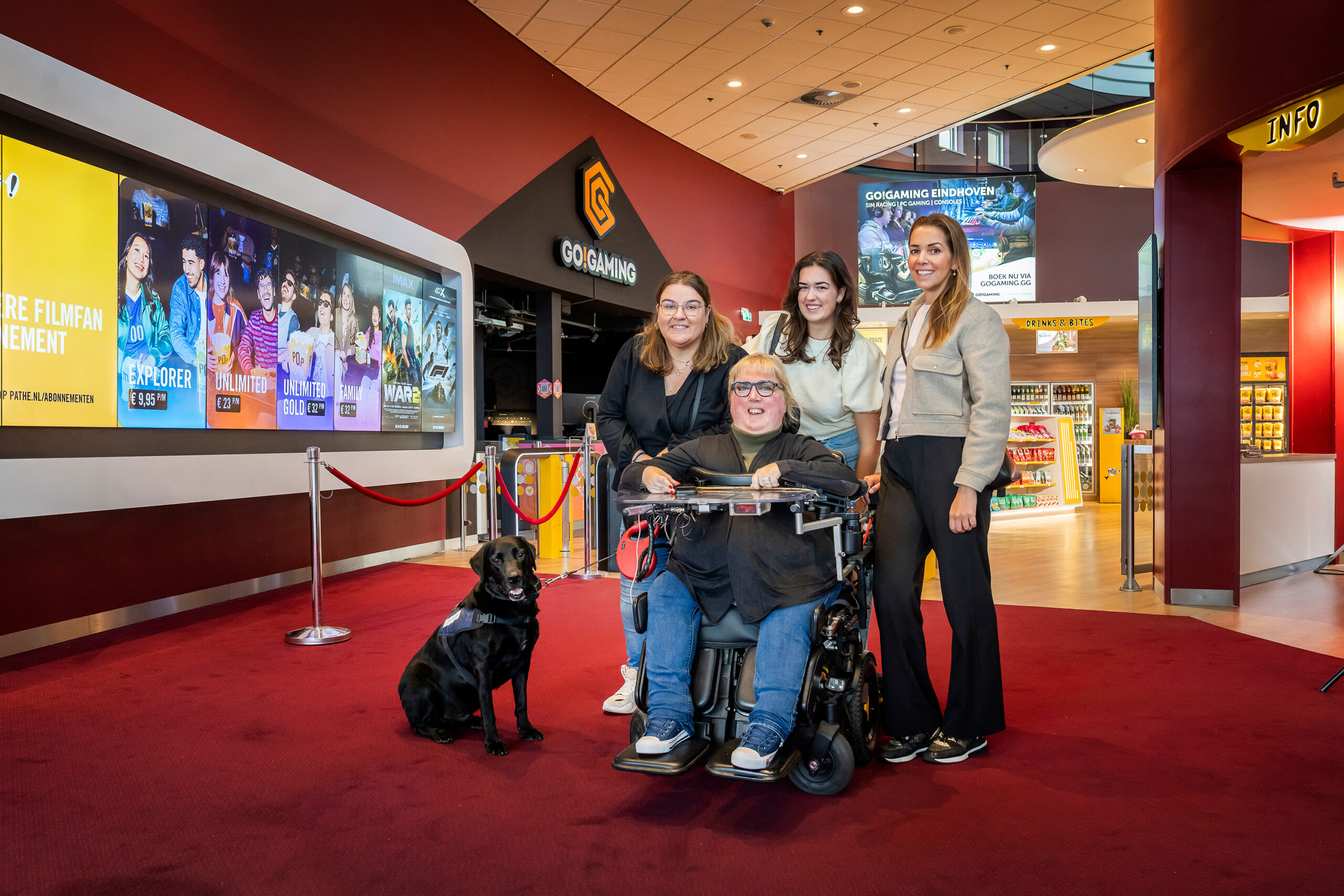 Marieke in haar rolstoel, haar vriendinnen staan achter haar. Iedereen lacht, haar hulphond zit naast de rolstoel